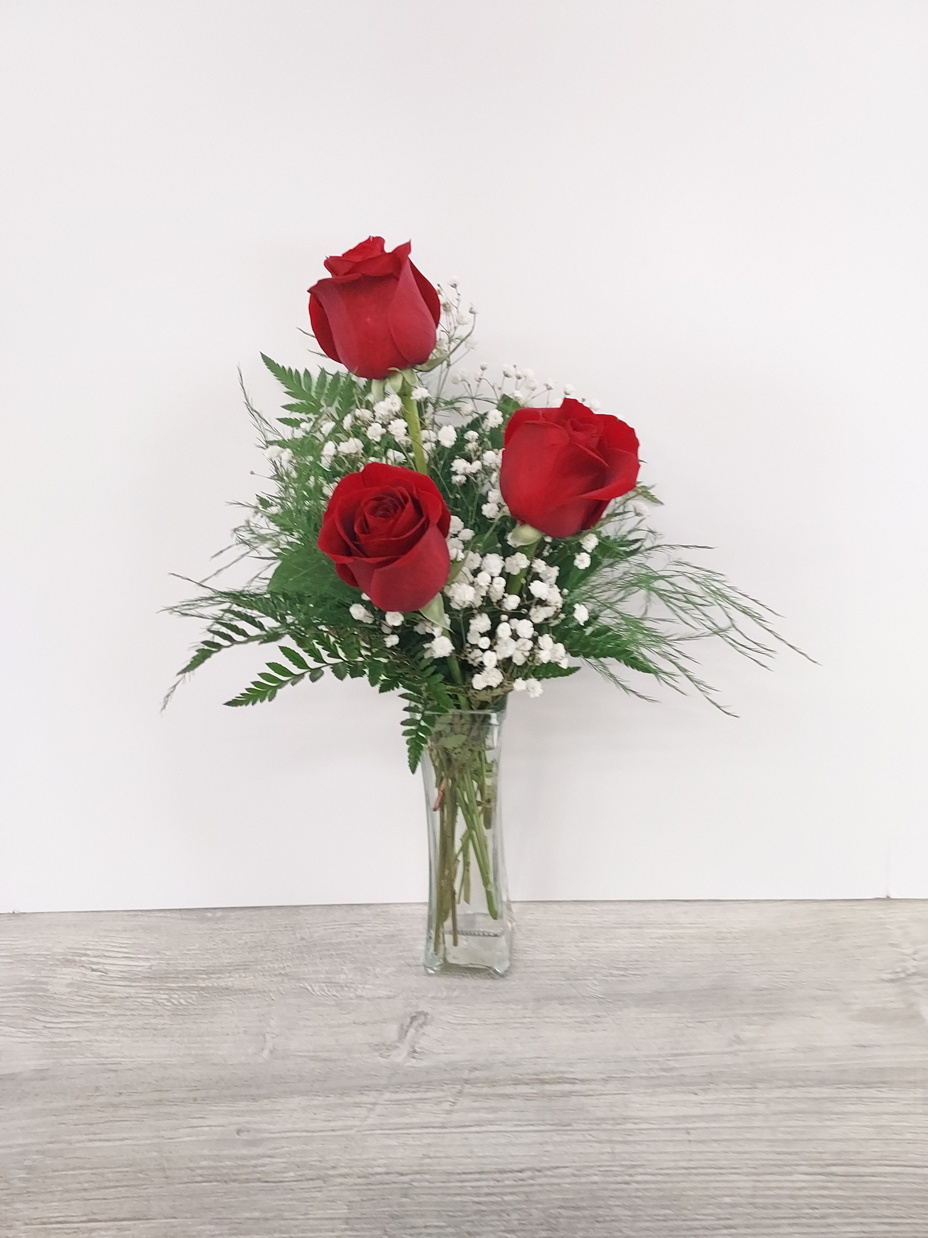 Tall clear vase with three red roses and small white accent flowers.