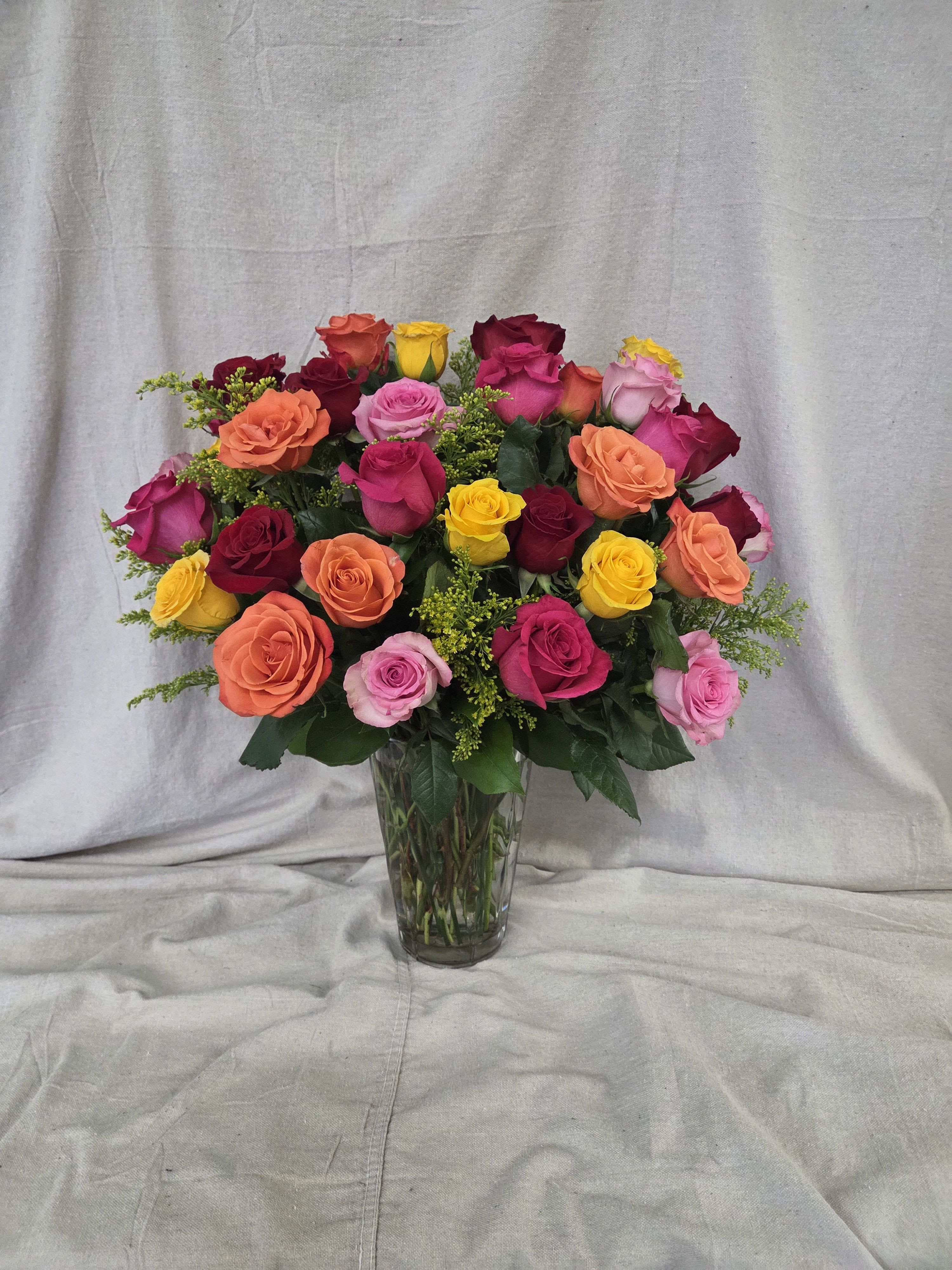 Tall clear vase filled with mixed red, pink, orange, and yellow roses with small yellow filler flowers