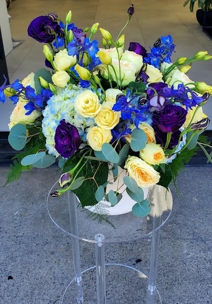 Low arrangement of yellow and cream roses, blue hydrangea, and purple blooms in a white vase on a clear stand
