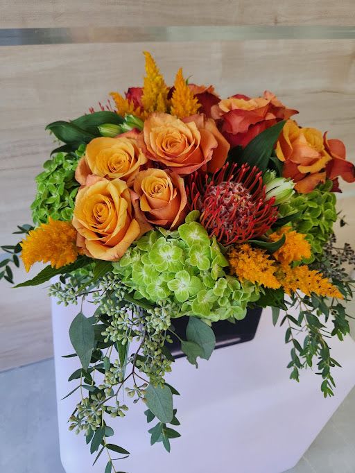 Low arrangement of orange roses, green hydrangeas, and red protea in a black container