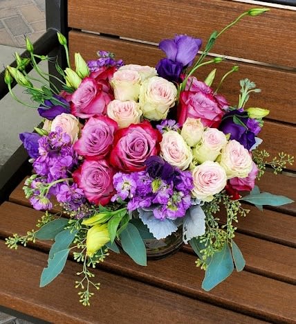 Low arrangement of pink and cream roses with purple flowers in a clear vase on a wooden bench