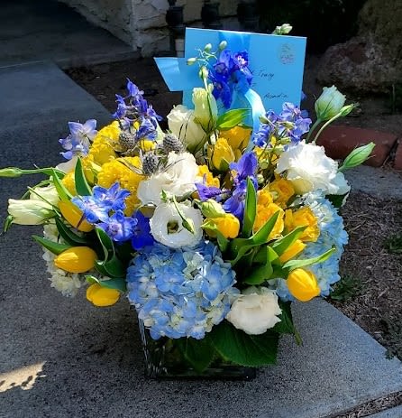 Blue, yellow, and white flower arrangement with tulips and hydrangeas in a clear glass cube vase and blue card