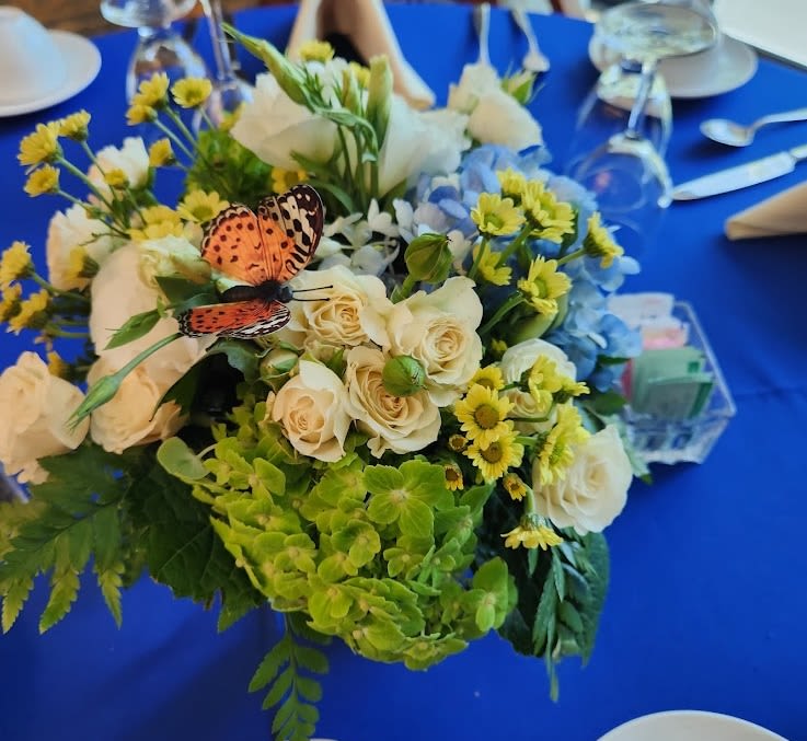 Low centerpiece of white roses, yellow daisies, and blue and green hydrangeas with an orange butterfly on a blue tablecloth