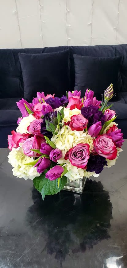 Low arrangement of pink and purple roses and tulips with white hydrangea in a clear glass cube vase
