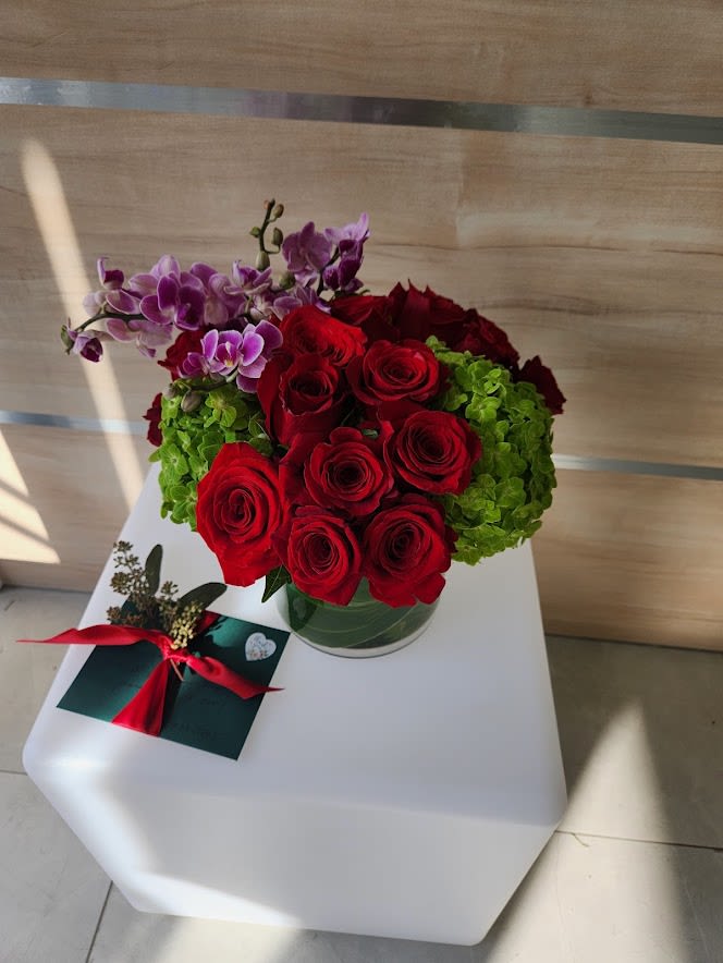 Low glass vase of red roses with green hydrangeas and purple orchids beside a ribbon-tied card