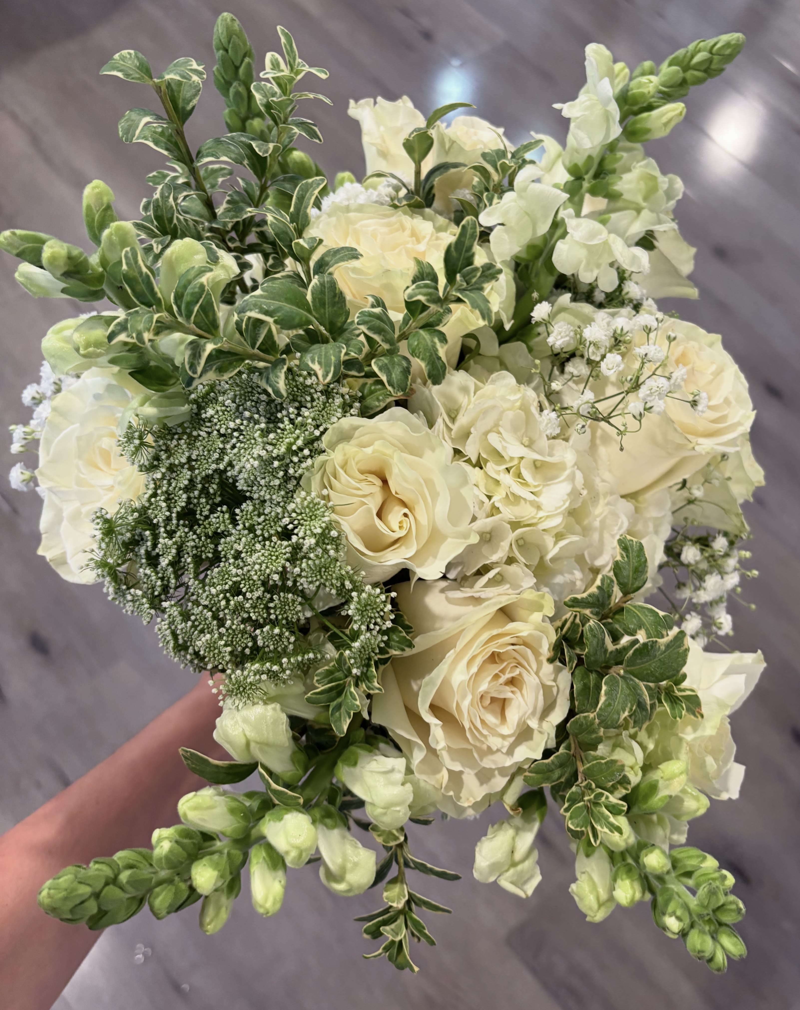 Cream roses and white flowers in a hand-held bouquet with variegated greenery.