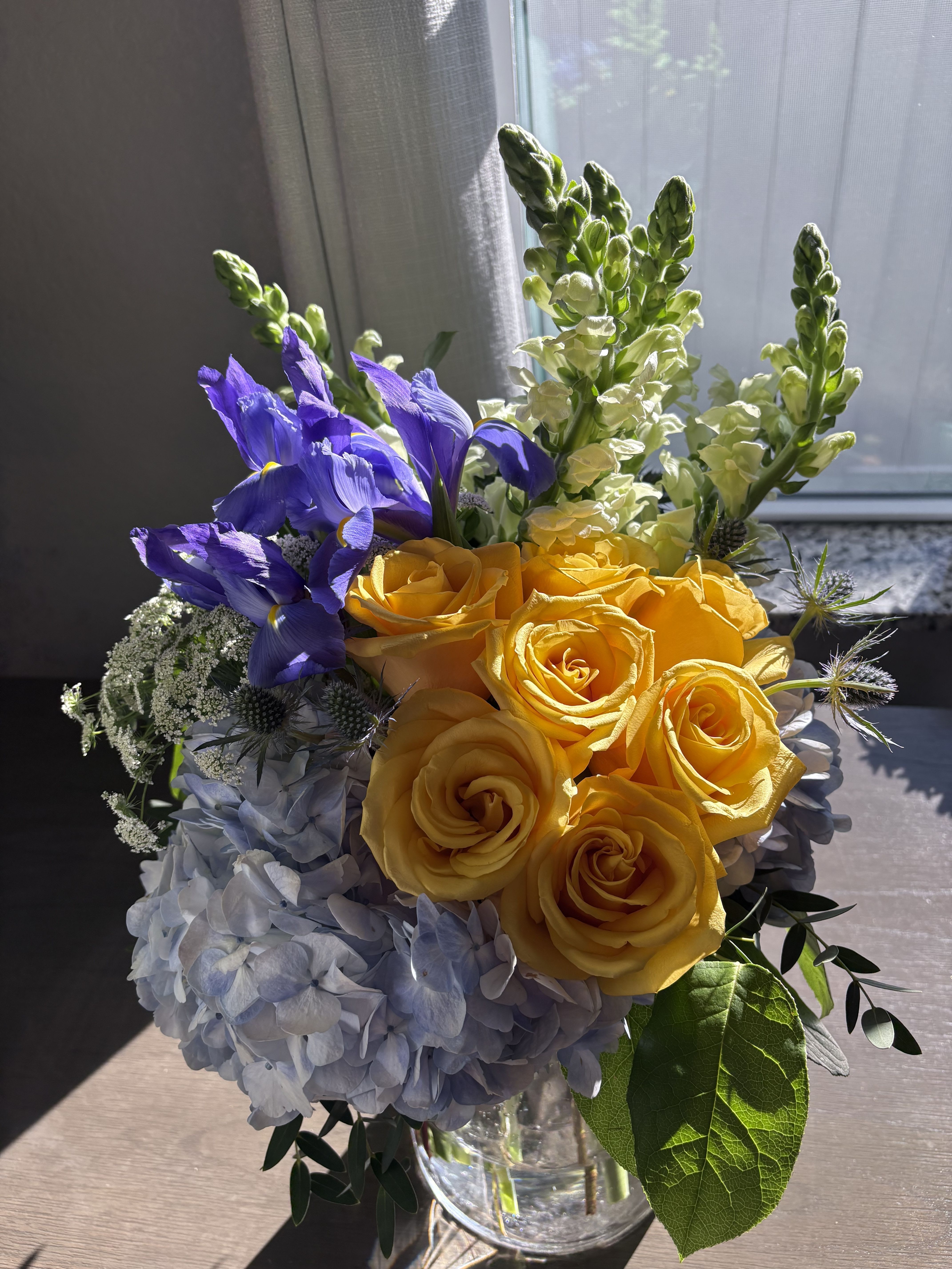 Bouquet of yellow roses, blue irises, and pale hydrangeas in a glass vase