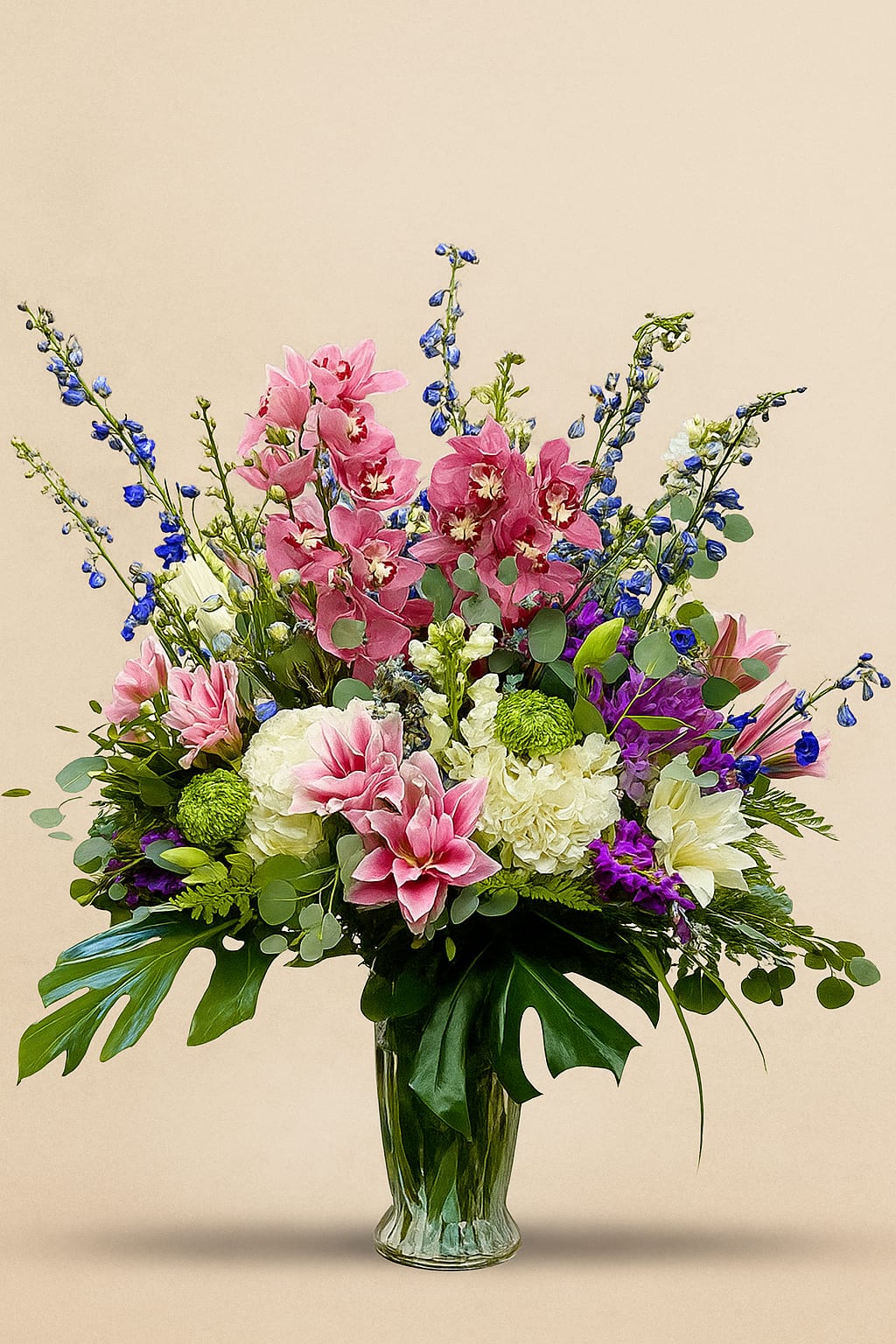 Grand Époque Arrangement - An opulent expression of floral artistry, our Grand Époque Arrangement evokes timeless elegance and refined grandeur. Towering stems of sapphire-blue delphinium and coral cymbidium orchids rise gracefully above a lush foundation of voluptuous hydrangea, creamy dahlias, and richly fragrant lilies. Accents of verdant foliage, including sculptural monstera leaves and silvered eucalyptus, lend dramatic texture and balance, while violet and chartreuse blooms provide unexpected notes of vibrancy.  Presented in a hand-polished glass vase, this arrangement is perfectly suited for a grand entry, formal dining setting, or any occasion demanding an unmistakable statement of sophistication.  At Savington &amp; Hill, every detail is a tribute to our heritage of bespoke floristry—crafted for those who appreciate beauty without compromise.