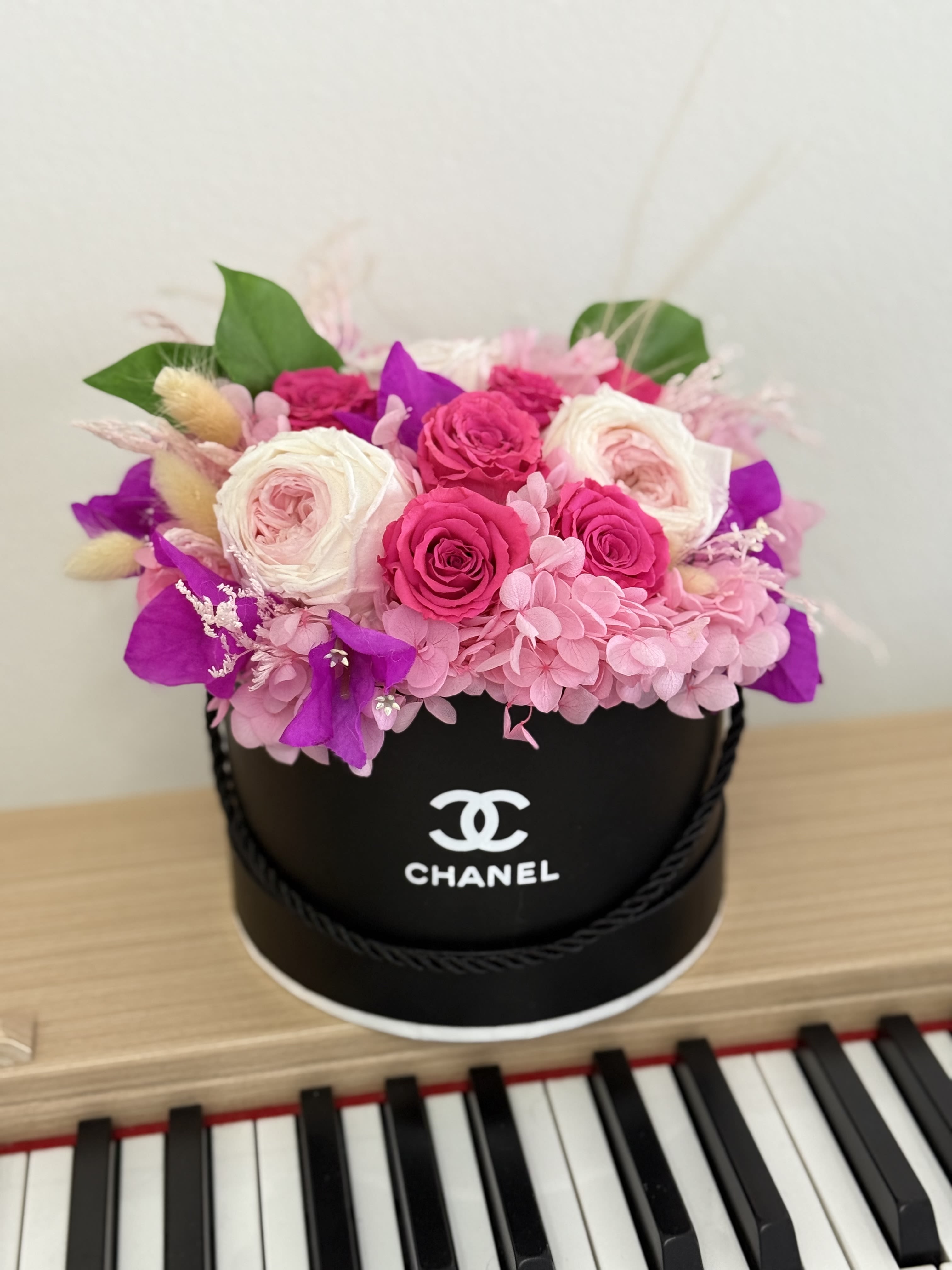 PInk Passion  - A lush cluster of tightly packed pink and white roses with pale pink hydrangea and vivid purple accents creates an immediate impression of romantic luxury - a birthday bouquet or anniversary arrangement that feels couture. Presented in a round black hat box with a printed logo and decorative ribbon, this floral gift reads as both fashion-forward and heartfelt; the bright pastels against the saturated magenta-purple blooms make it a joyful choice for celebrations from graduation and prom to Mother's Day or just because.  Silk roses and hydrangeas are arranged densely for a polished, long-lasting look that suits Thanksgiving centerpieces, Christmas gifting, housewarming presents, or a thoughtful get-well or thinking-of-you surprise. The black hat box adds modern contrast and makes the arrangement easy to display on a mantel, bedside table, or entry console; dimensions follow standard hat-box sizing for compact tabletop presence. The preserved silk construction means the arrangement requires no watering and keeps its fresh appearance for years, making it an enduring reminder for birthdays, anniversaries, new baby greetings, or holiday celebrations like Valentine's Day and Hanukkah.