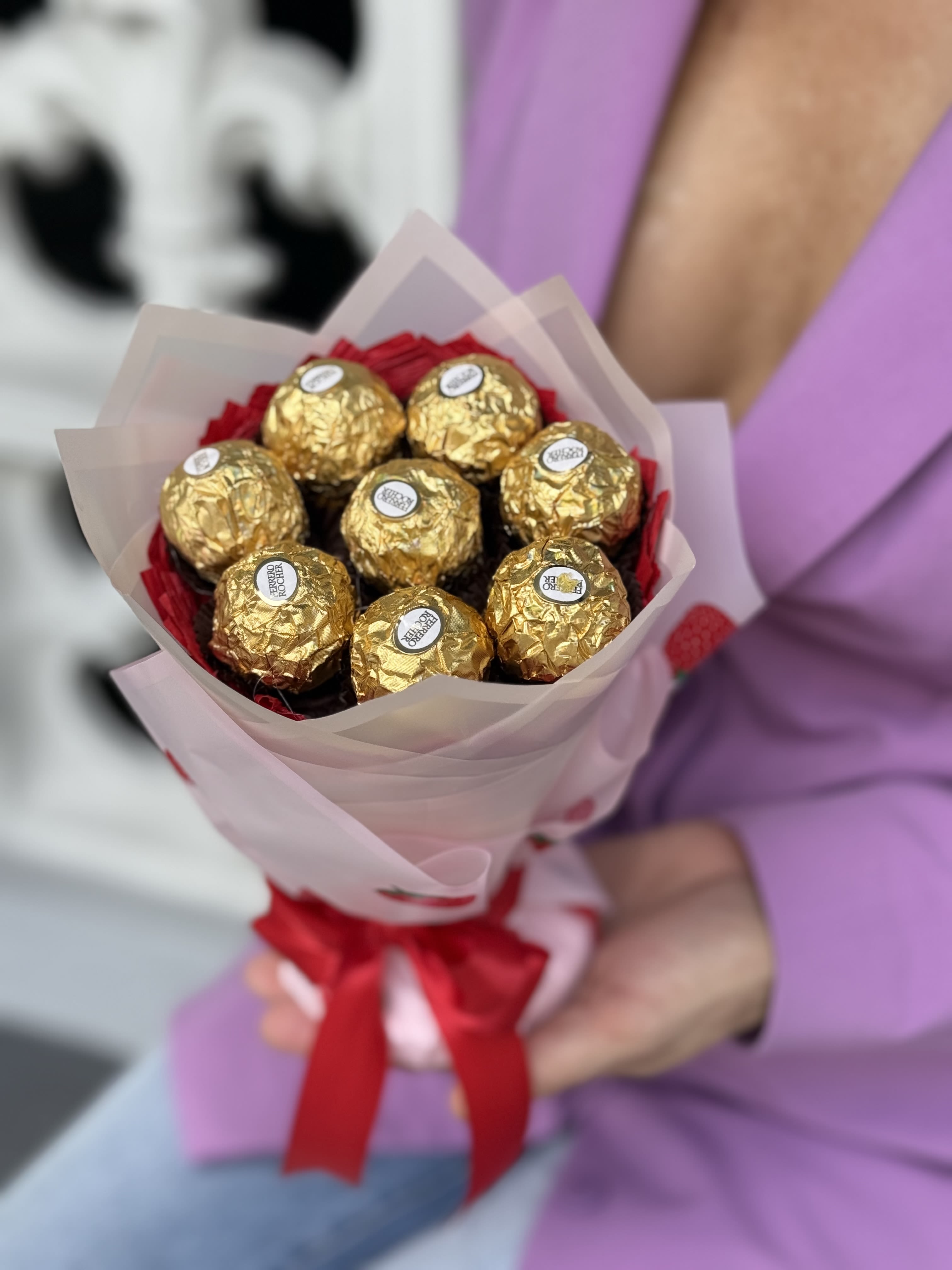 Golden Delight Ferrero Rocher Bouquet - Glistening gold Ferrero Rocher chocolates artfully arranged like a hand-tied bouquet give this chocolate bouquet a festive, romantic presence perfect for anniversary roses, birthdays, Valentine's Day, or a sweet "thinking of you" gift. The metallic gold clusters catch the light against soft wraps of pale pink or translucent paper and a bold red satin ribbon, so the recipient will feel celebrated the moment they see it - a confectionary alternative that reads like a floral tribute.  Each arrangement is crafted from gold-foil wrapped Ferrero Rocher candies layered in white or pale pink wrapping paper with red accents and playful strawberry prints, then finished with a bright red satin ribbon for a polished, gift-ready look. The palette of gold, red, pink, white and a touch of yellow creates a warm, seasonal feel that suits Thanksgiving, Christmas, Mother's Day, graduations, housewarmings, get-well wishes, or just-because surprises. Sized like a traditional hand bouquet, it's easy to carry, display, and share; no vase needed and no special care - simply present and enjoy.