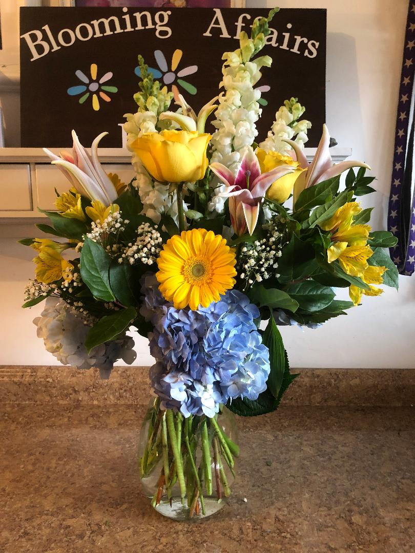 Mixed bouquet with yellow lilies, blue hydrangeas, and a yellow gerbera daisy in a glass vase