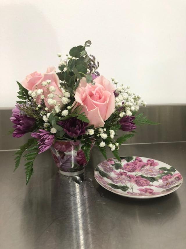 Pink roses and purple flowers in a clear vase beside a floral plate