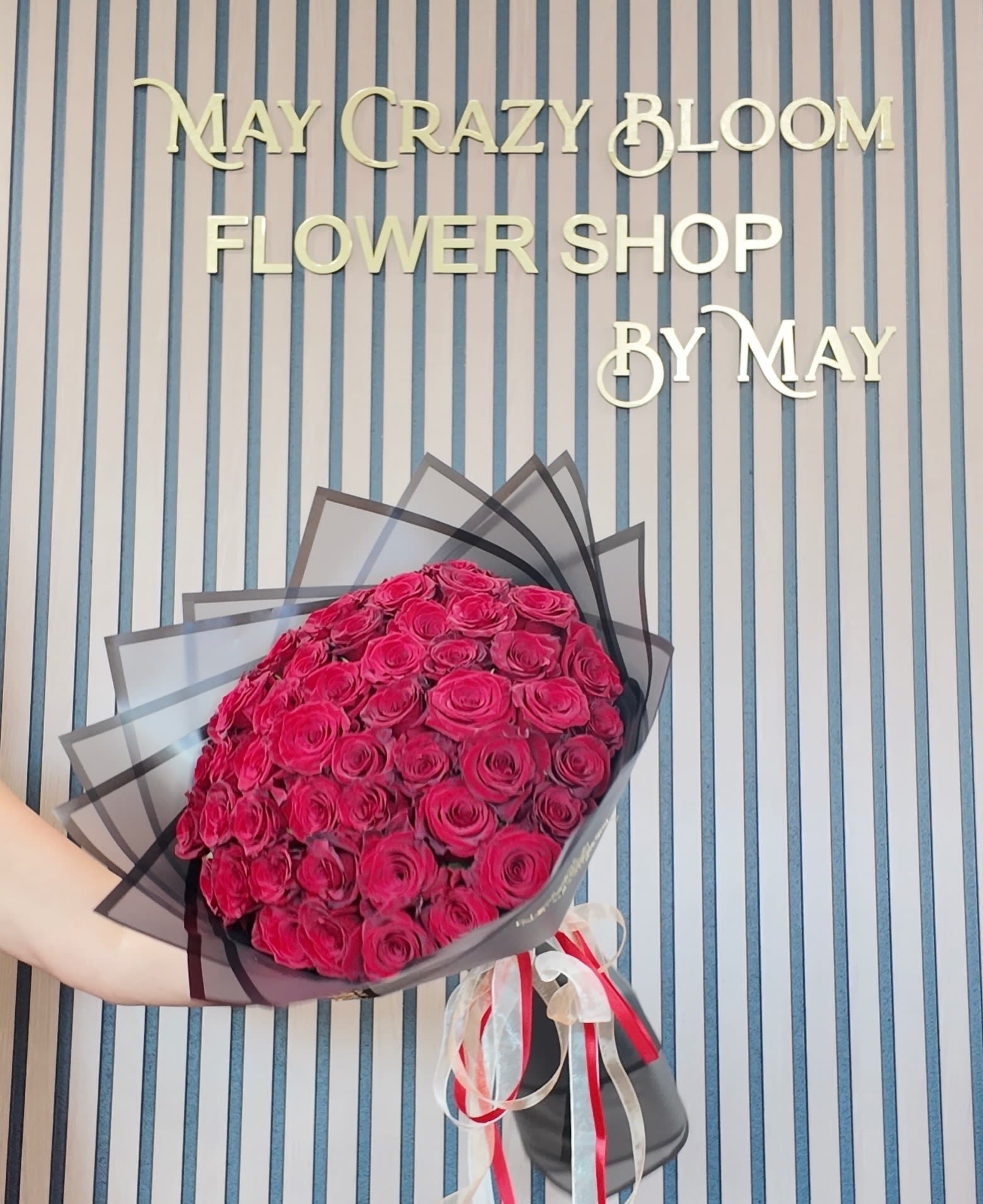 36 red roses - A lush dome of 36 tightly packed roses in saturated pink-red tones creates an immediate wow - the kind of birthday or anniversary bouquet that makes the person you're thinking of feel adored. This handheld arrangement reads romantic and celebratory, equally right for saying "get well soon," "congrats," or "thinking of you" when words fall short.  Each rose is arranged in a compact, rounded hand-tied shape and wrapped in layered black and dark gray paper for dramatic contrast. The bouquet is finished with a sheer wrap and twin ribbon accents in red and white, giving a polished, gift-ready presentation that also suits graduations, Mother's Day, weddings, or a thoughtful "I'm sorry." The small, dense silhouette makes it easy to carry or place in a vase at home; to keep the blossoms at their best recut stems and change the water every other day.