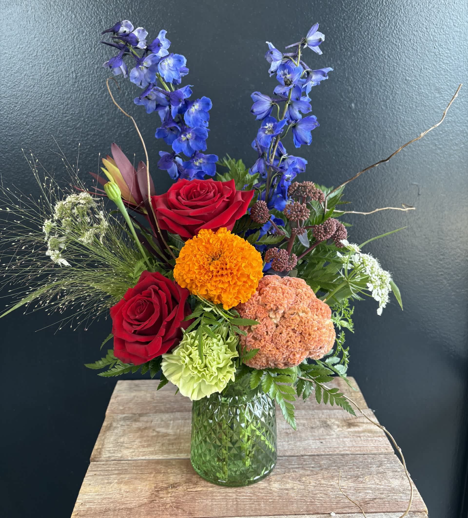 Mixed bouquet with red roses, blue delphinium, and orange blooms in a green glass vase