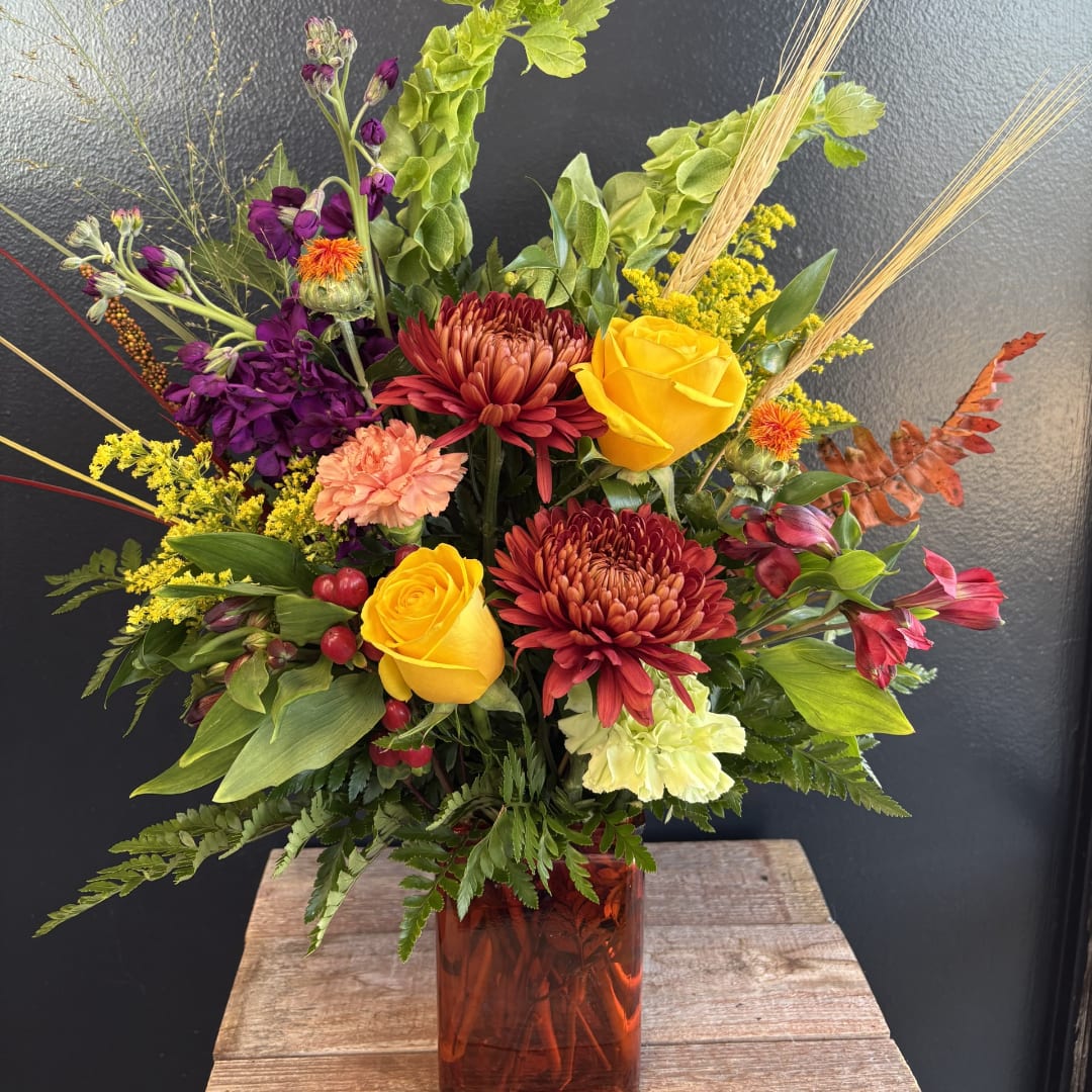 Mixed bouquet with yellow roses, burgundy mums, and purple flowers in an amber vase