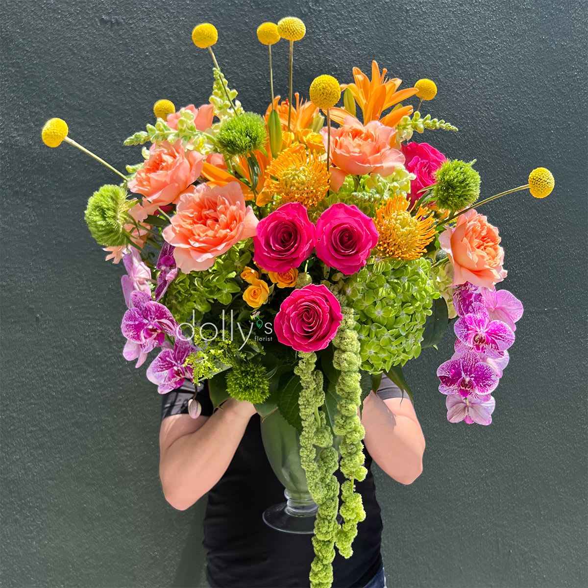Citrus Opulence - A lush centerpiece that tastes of sunshine and zest - Citrus Opulence is a large flower arrangement perfect for a cheerful birthday bouquet, a bright anniversary surprise, or any moment you want to make someone feel deliciously celebrated. The design centers on unique grapefruit-colored roses paired with hot pink roses and peach carnations, while purple orchid sprays add an unexpected luxe note; the overall effect reads fresh, vibrant, and joyfully abundant for gifting or bold everyday display.  This exuberant mixed bouquet sits in a clear glass vase and features orange accent blooms, chrysanthemum touches, and bright yellow spherical stems that rise above the dense center to give the composition a playful, sculptural silhouette. A long trailing green element spills down from the vase, reinforcing the cool green backdrop and providing lively contrast to the warm citrus palette. Sized as a large arrangement, it fills a room with fresh color and texture; to keep it looking its best, refresh the water every couple of days and trim stems as needed when rearranging.