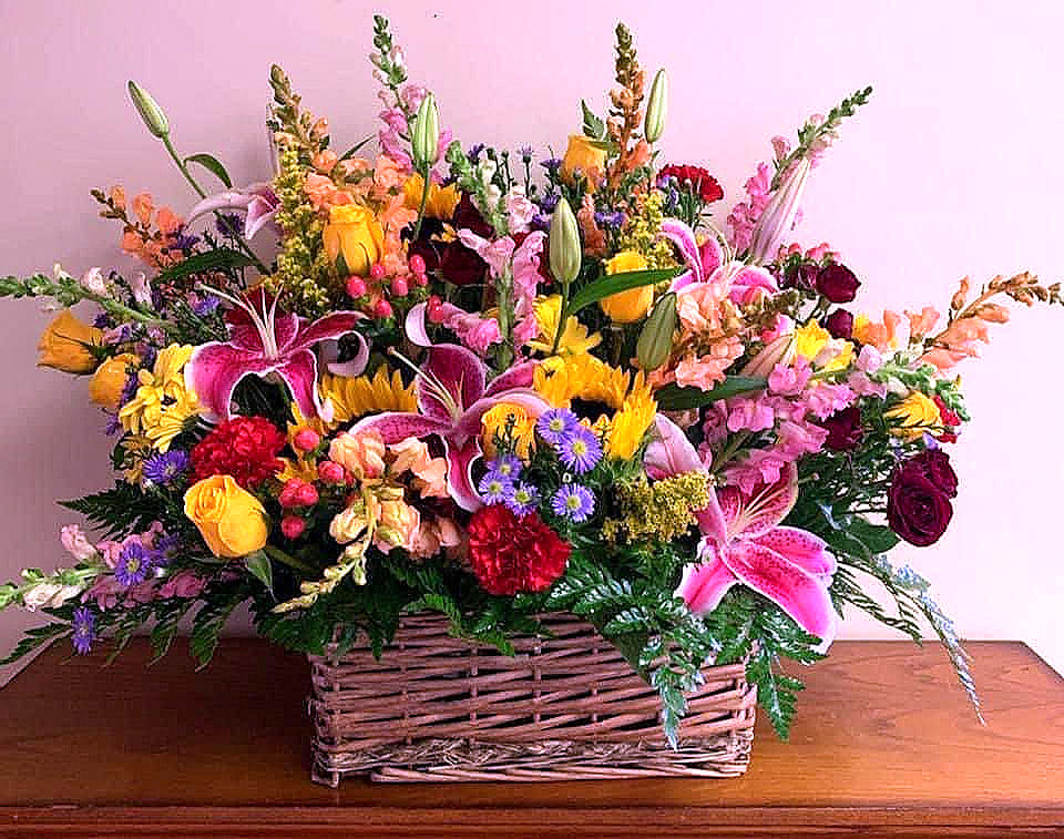 Large mixed basket arrangement with pink lilies, yellow roses, sunflowers, asters, and red carnations.