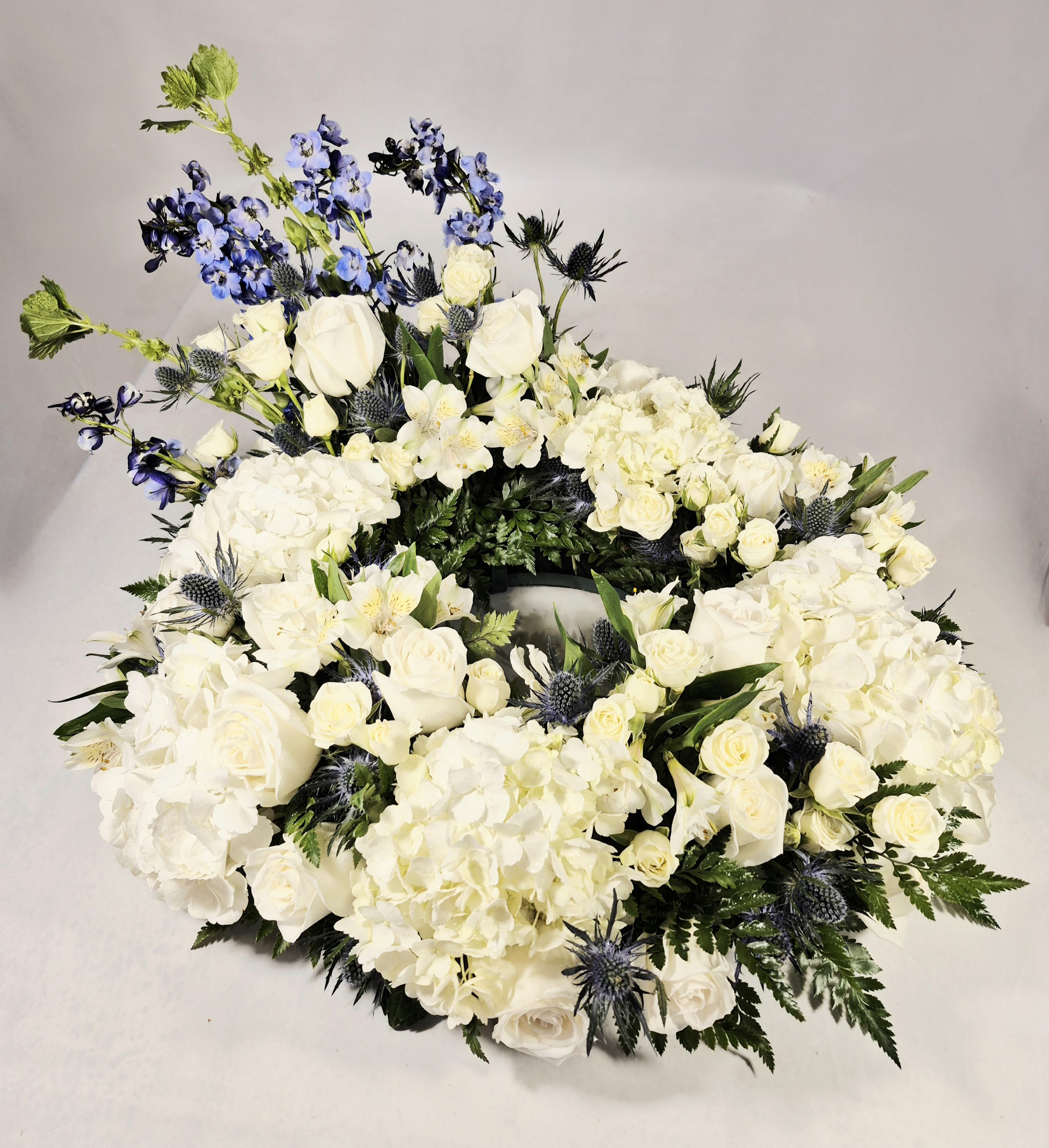 White Hydrangea and White Rose Urn Wreath - A serene circular wreath of white hydrangea and white roses creates a calm, reverent focal point for sympathy and funeral tributes. This white floral urn wreath conveys quiet elegance and respect-the kind of arrangement that helps the person you're thinking of feel honored and gently remembered at a memorial service or graveside.  Densely packed hydrangeas, full roses, spray roses, and alstroemeria form the soft, textured ring, while tall blue delphinium stems and spiky blue thistles rise from one side to add cool contrast and vertical interest. The wreath sits on a sturdy wreath base with an open center and a formal, structured silhouette; the white, blue, and green palette reads peaceful and composed from every angle.  Designed for sympathy use but suitable year-round, this arrangement offers a timeless expression of comfort for family and friends. 