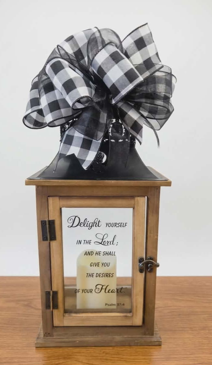 Delight Yourself in the Lord Lighted Lantern - A rustic wooden lantern with a black metal roof and a bold black-and-white buffalo check bow offers a warm, faith-centered gift for someone who cherishes inspirational home décor. This faith-based decorative lantern is ideal as a sympathy gift, encouragement present, or thoughtful housewarming accent that quietly celebrates devotion and comfort.  The lantern features clear glass panels and a metal latch; the front glass displays a printed Bible verse for daily encouragement. Finished in a natural, slightly weathered wood tone and topped with a large check ribbon, it brings a simple farmhouse look-monochrome, timeless, and easy to place on a mantel, entry table, or bedside.  Make this the piece you send to let someone know you're thinking of them-perfect for a friend, family member, or church leader who would appreciate a meaningful, long-lasting keepsake. The lantern arrives ready to display and pairs beautifully with a small battery operated candle (batteries included) to softly illuminate the verse. Lantern Reads " Delight yourself in the Lord; and he shall give you the desires of your Heart. Psalm 37:4" 20" T 5 1/2" D 8 1/2" W