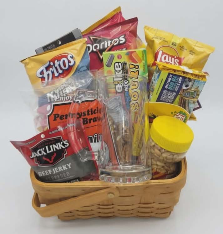 Snack Basket w/ Mug  - Bright, casual, and made for sharing - this snack basket with a mug is a cheerful gift basket for who prefers salty, sweet, and ready-to-eat treats over flowers. Perfect for a casual celebration, or an office thank-you, the assortment reads as a low-fuss, feel-good present that says "you're appreciated" in a practical, tasty way the person you're thinking of can enjoy right away.  A woven basket holds an upright assortment of chips, jerky, candy, and other packaged snacks in bright yellow, red, and multicolored packaging, with a clear plastic snack container and a small plastic jar of nuts (yellow lid) arranged front and center. The included mug tucks into the mix for a usable keepsake, and the compact, grab-and-go presentation makes it easy to place on a desk or snack table. Ideal for the dad, coworker, or friend who loves variety, order to ensure same-week delivery for holidays and gatherings; no special care is needed beyond sharing the snacks. Comes wrapped in clear cellophane and bow. 