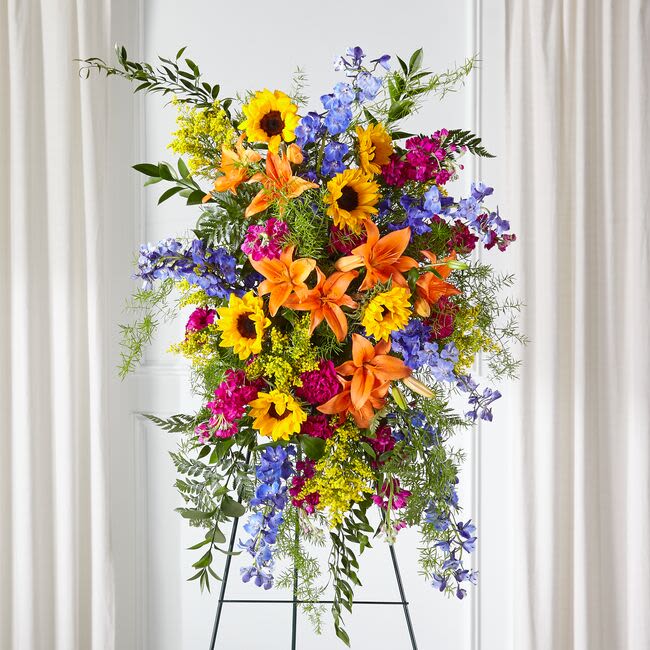 Tall standing spray of sunflowers, lilies, and blue flowers on an easel