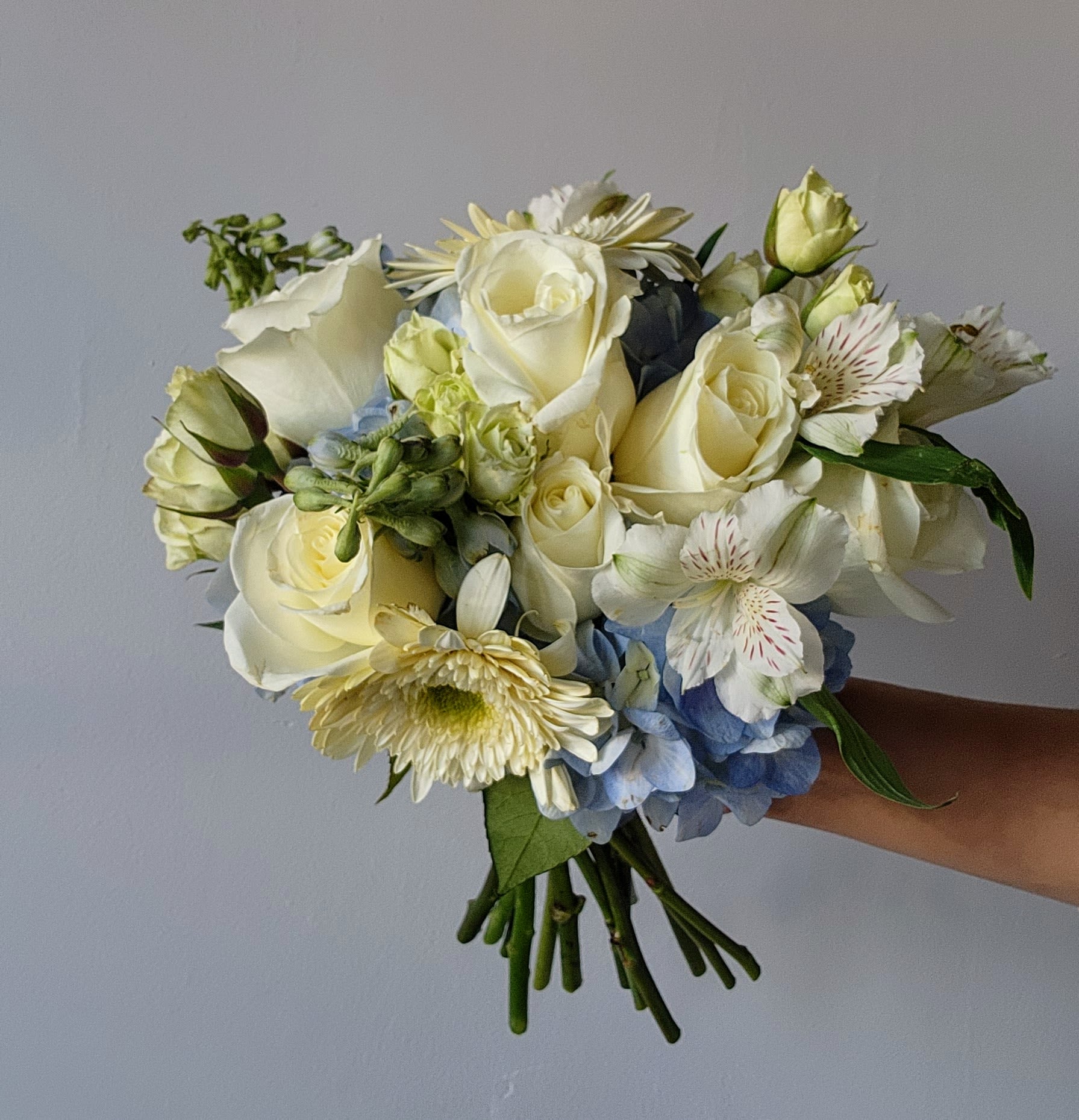 Handheld bouquet of white roses, blue hydrangea, and pale daisies