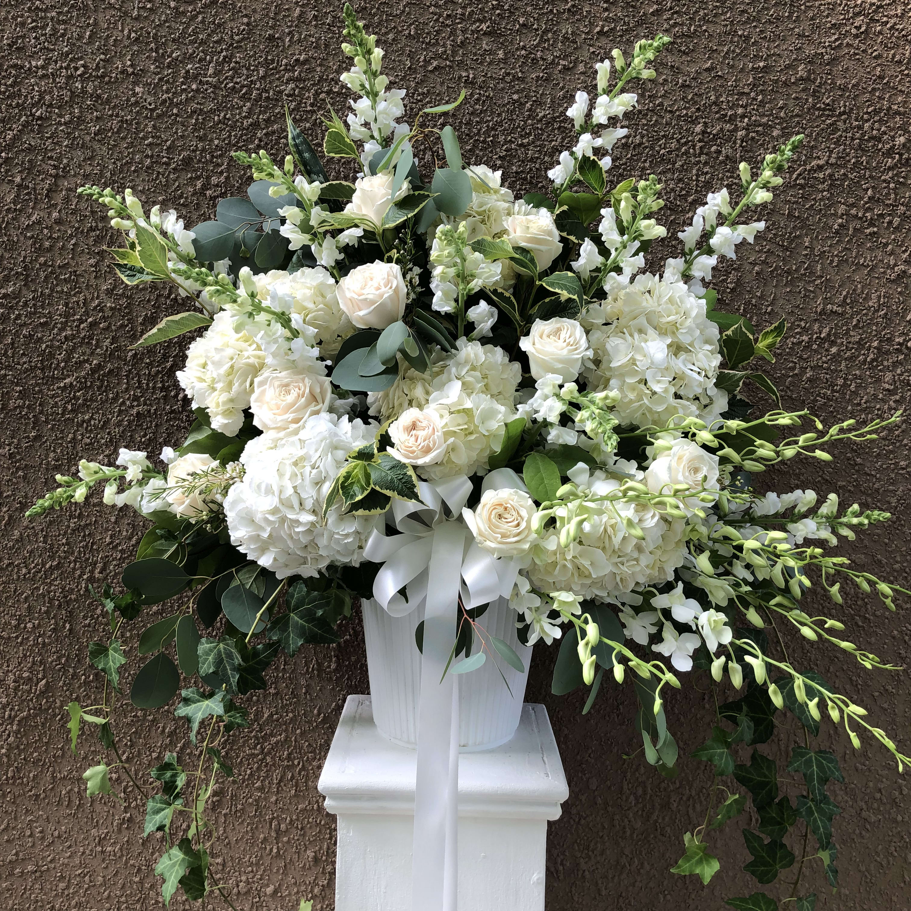 White floral arrangement in a white pedestal vase with a ribbon bow