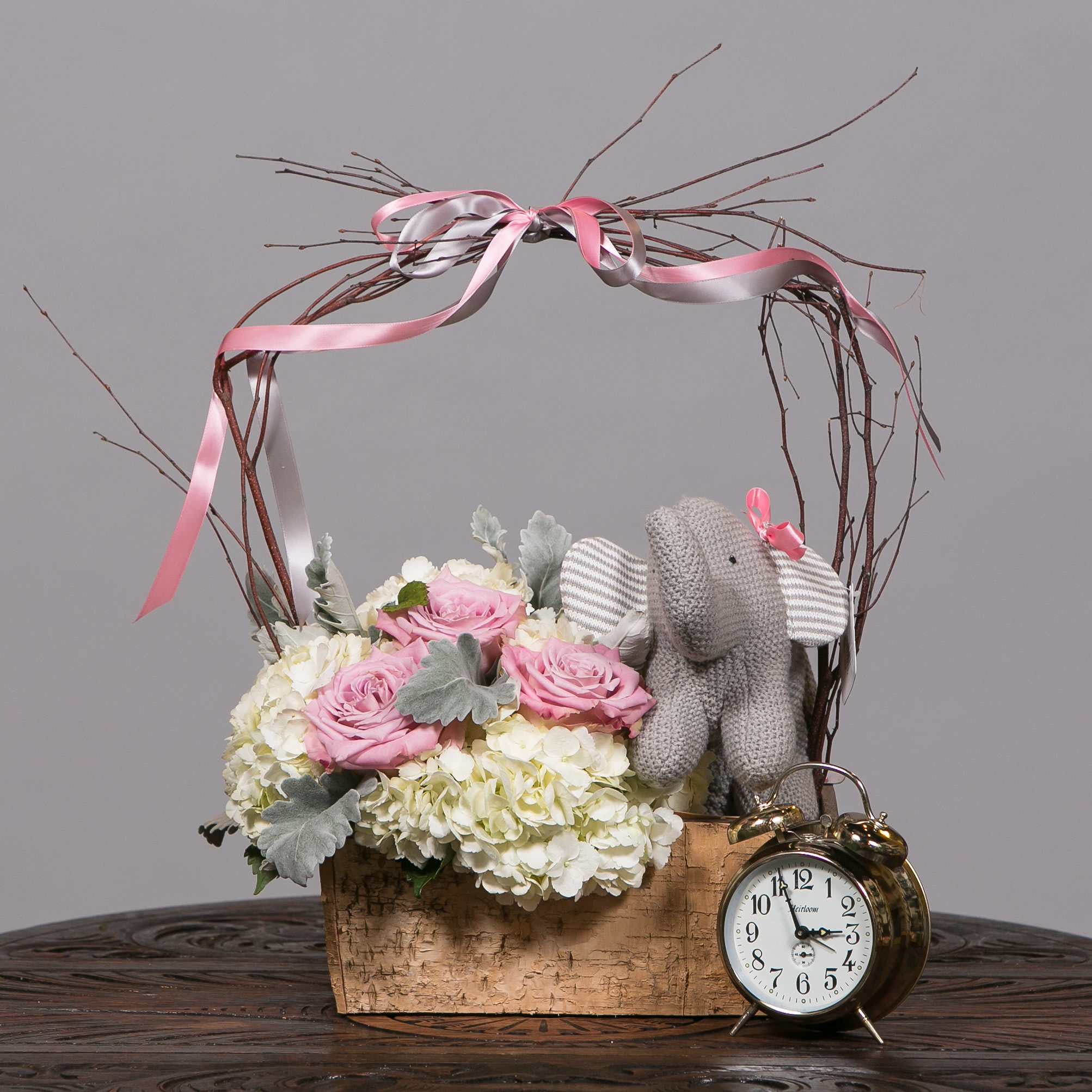 Pink roses and white hydrangeas in a wooden basket with a knitted elephant and clock