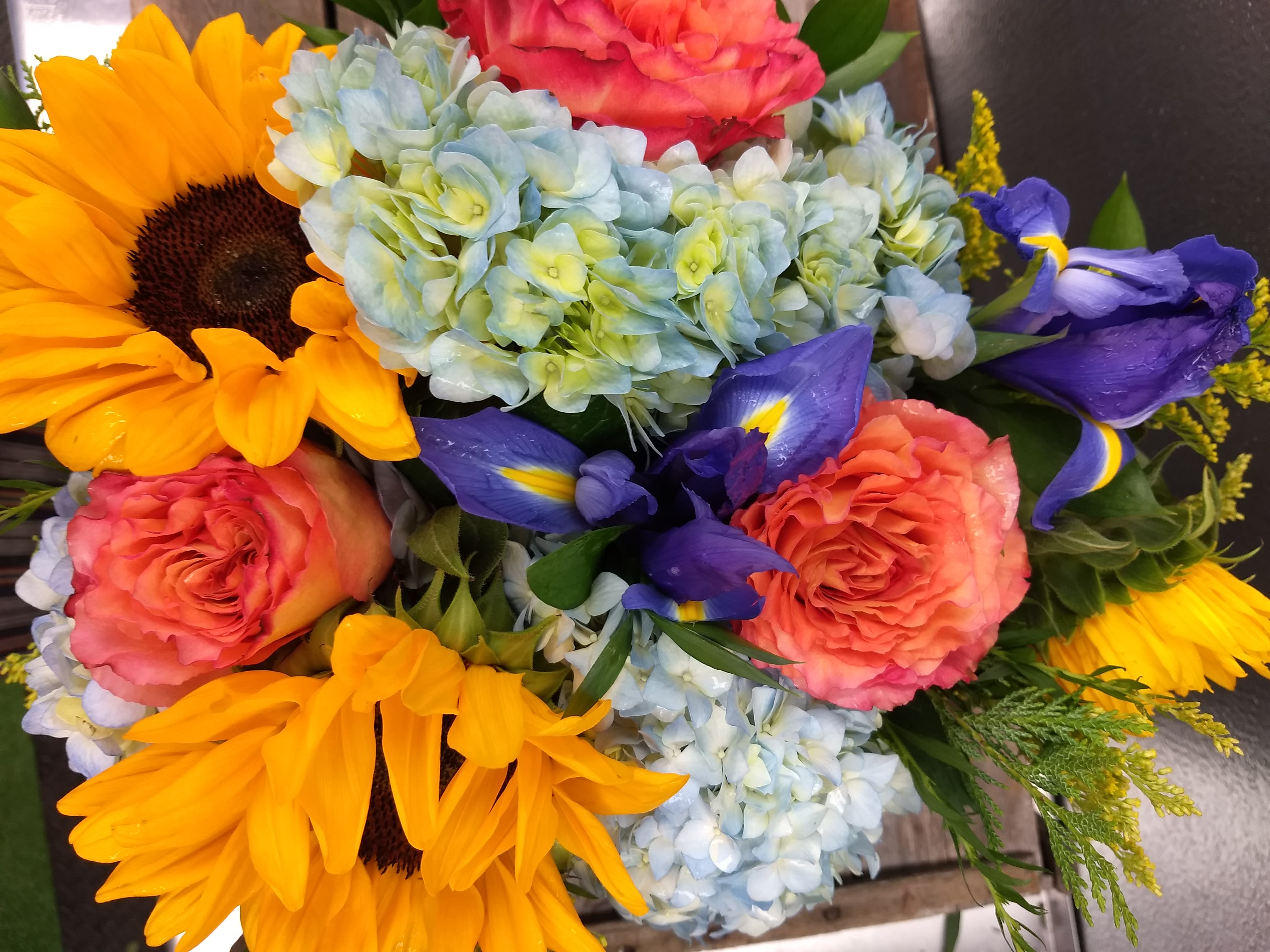 The Munroe - Big golden sunflowers take center stage in this cheerful flower arrangement, making The Munroe a perfect birthday bouquet, get well gift.  This lush, low design pairs large sunflowers with ruffled  bi colored orange roses, pale blue hydrangea clusters, and deep irises for striking color contrast and varied textures. Fine yellow filler blooms and airy greenery frame the composition for a full, garden-fresh look; the flowers are closely clustered to create a generous, abundant presentation that makes the recipient feel truly seen and celebrated.  Whether you're brightening someone's day "just because," congratulating a loved one, or adding warm color to a table. 