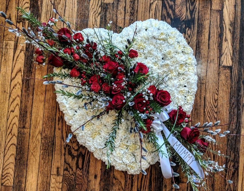 Heart-shaped white funeral arrangement with red rose spray and 'MUMMY' ribbon on a wooden floor