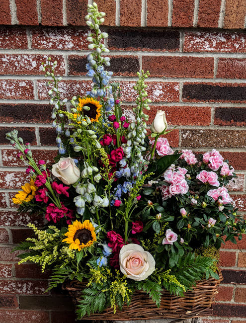 Tall mixed arrangement with sunflowers, roses, blue delphinium, and pink flowering plant in a wicker basket.