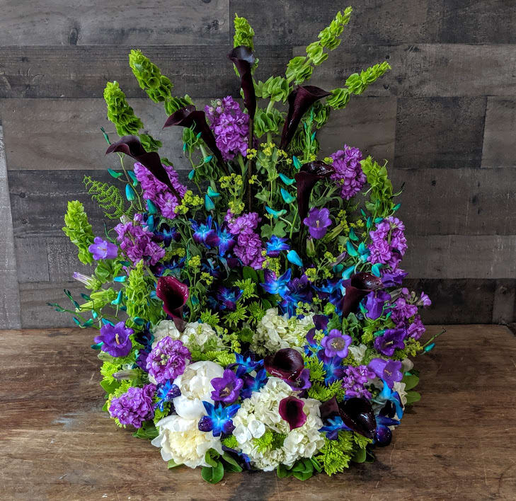 Tall one-sided arrangement with purple blooms, blue orchids, dark calla lilies, and white hydrangeas