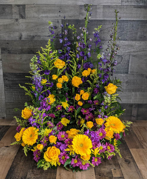Tall one-sided arrangement of yellow roses and gerbera daisies with purple delphinium and daisy mums