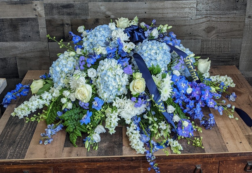 Low blue and white casket spray with hydrangeas, roses, and delphinium accented by navy ribbon.