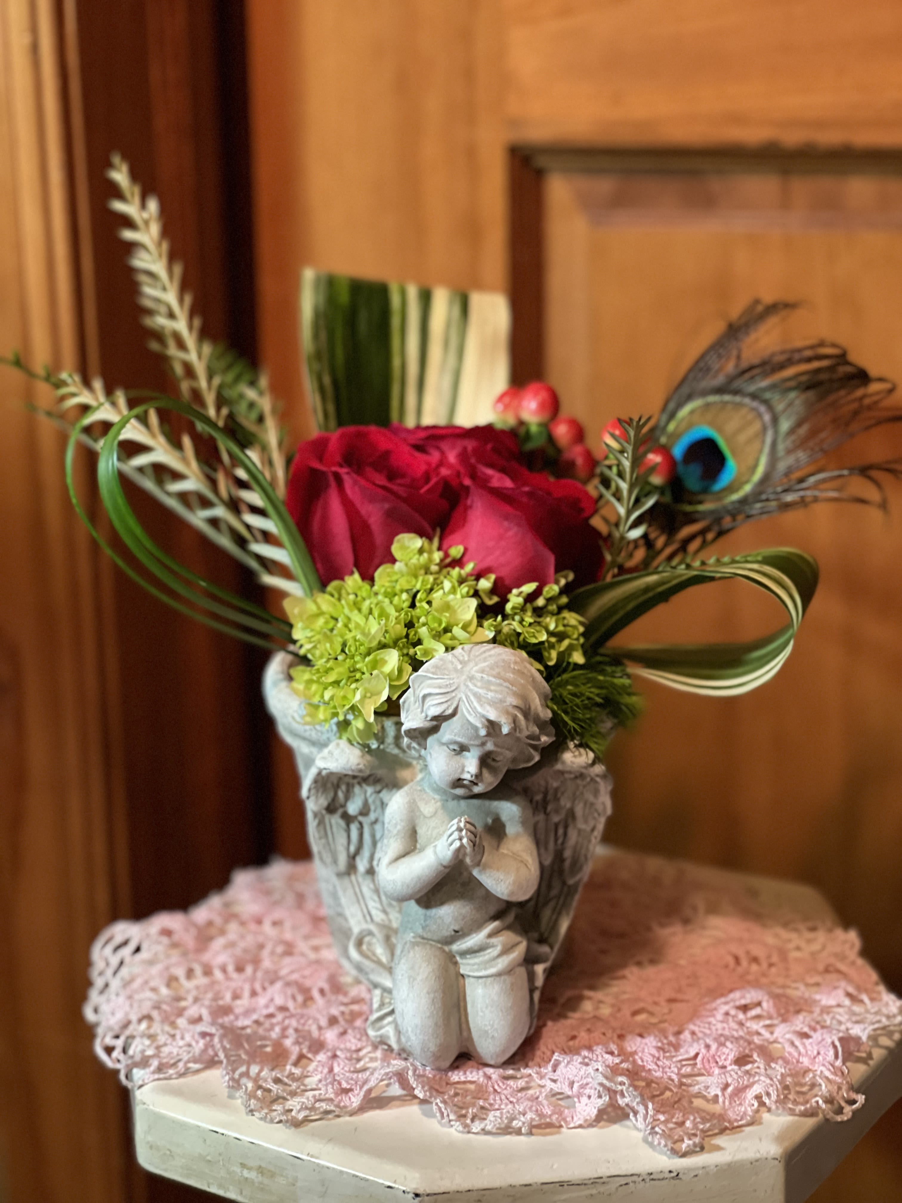 Red roses and green blooms in an angel vase with looped foliage and a peacock feather accent.