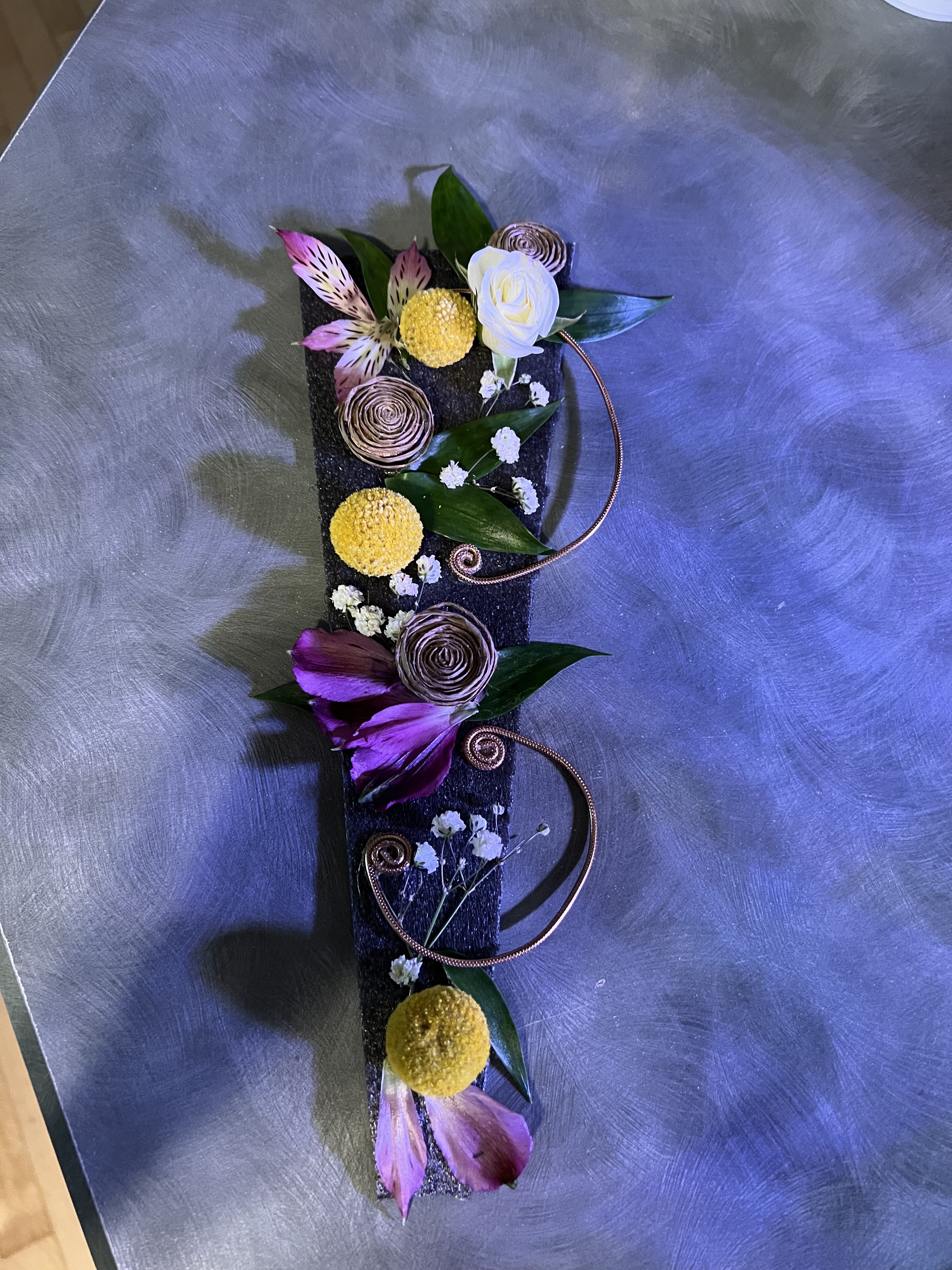 Vertical floral arrangement on a dark rectangular base with purple and yellow blooms