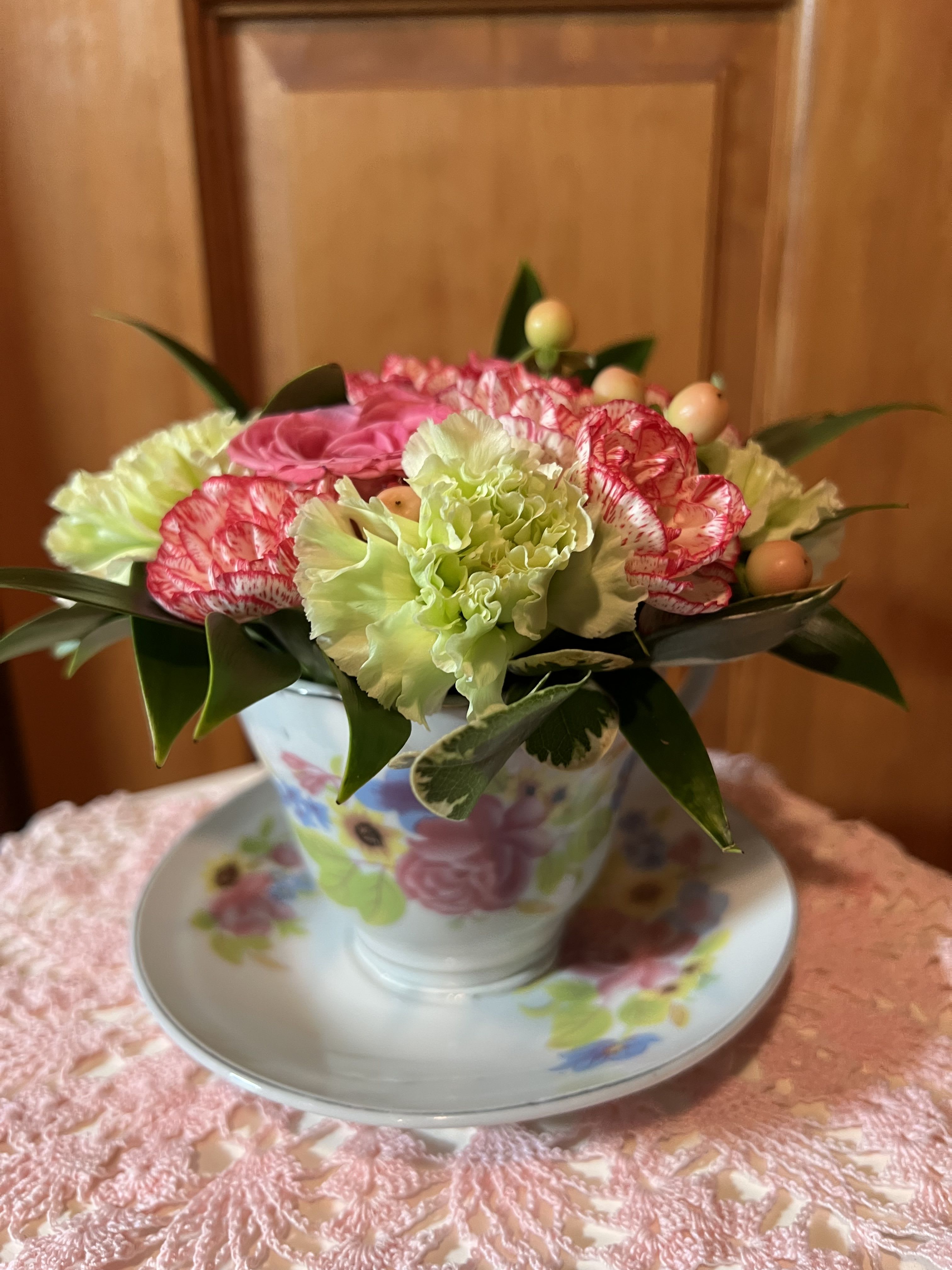 Small arrangement of pink roses and green carnations in a floral teacup with saucer on a lace cloth