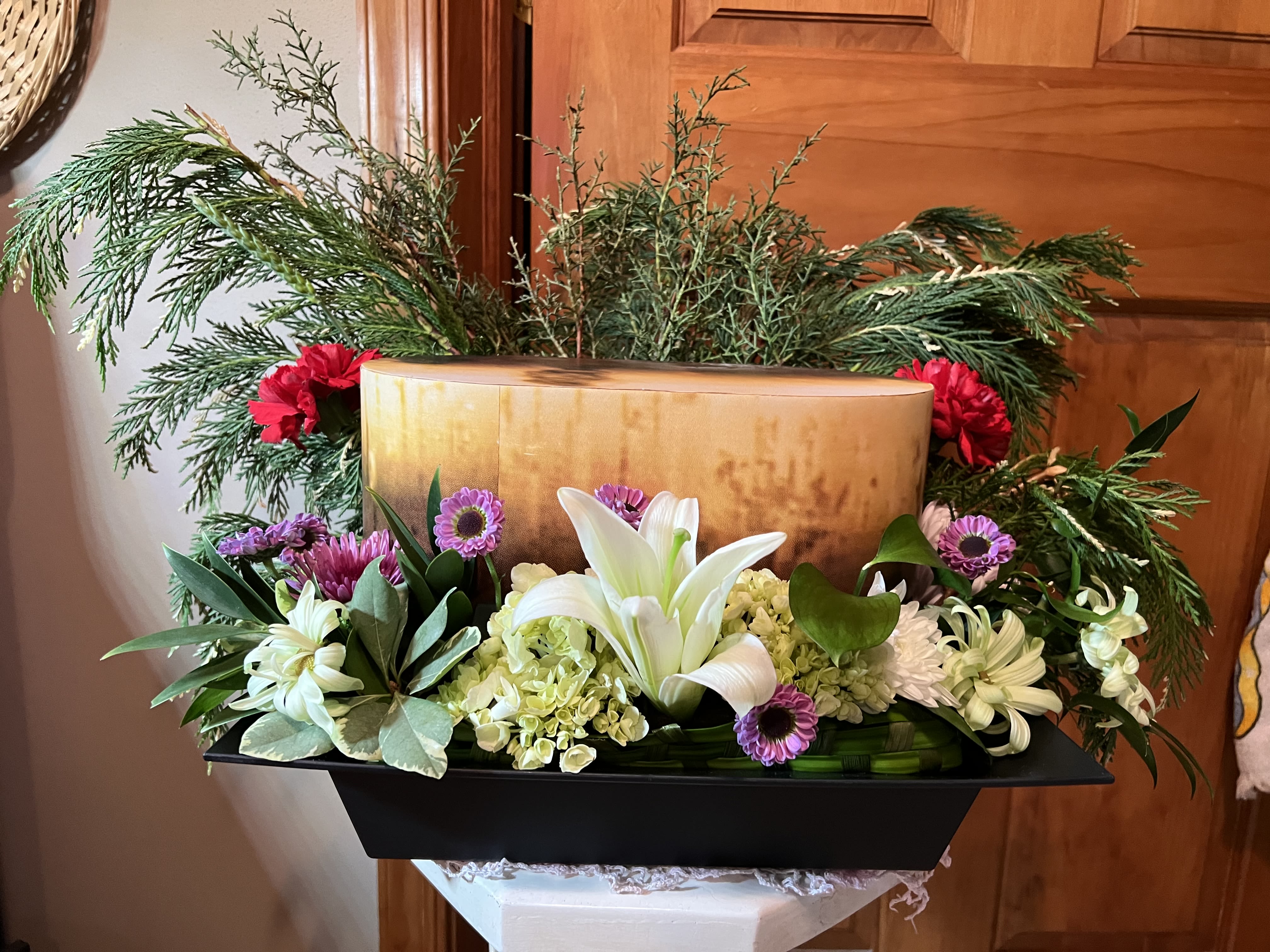 (NEW) Rectangular Urn Cremation Hug - A low, dignified rectangular cremation arrangement that feels both elegant and quietly celebratory-perfect for sympathy services, memorials, or a winter tribute. White lilies anchor the center to convey reverence, while touches of red and purple bring warmth and depth so the person you're honoring looks and feels truly remembered.  This large design is set in a sleek black container, giving it a formal, grounded presence at a service or home memorial. Pale green hydrangea clusters and purple daisy-like blooms layer around the lilies, with bright red accents and textured evergreen branches framing the composition for fullness and seasonal contrast. The mix of chrysanthemums and other filler blooms adds variety in shape and long-lasting structure.  Made to sit low and broad, this piece reads well at the front of a service table or on a memorial display, offering a composed, substantial tribute that honors the recipient without overwhelming the space. Care is straightforward-kept cool and refreshed with water at the stand-to preserve the lilies and hydrangeas for the days they're most needed.