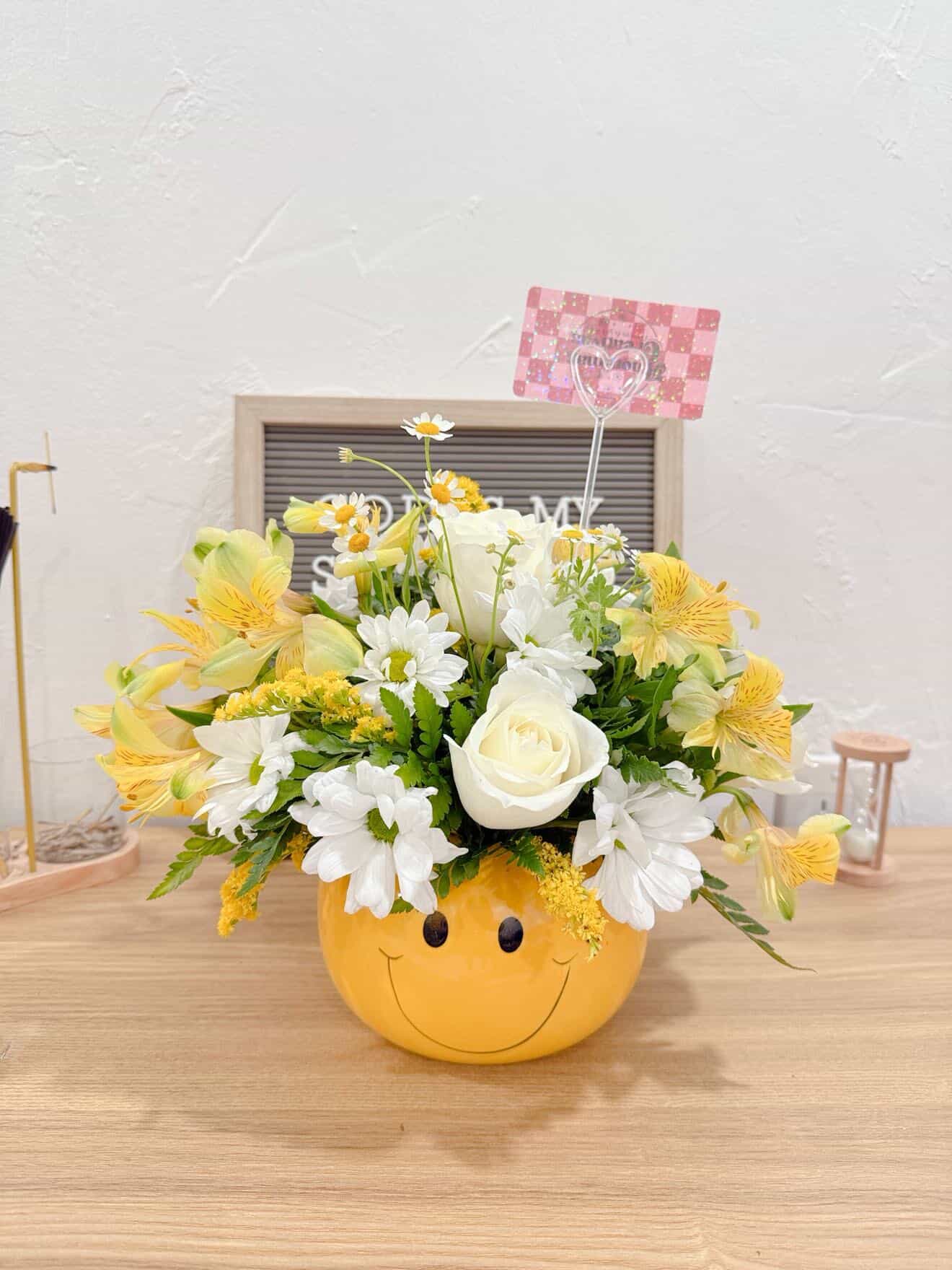 smile -  Smile Bouquet – A Gift That Brightens the Heart   Bring joy to someone special with this beautiful floral arrangement presented in a charming yellow smiley-face container. Designed with elegant white roses, delicate daisies, yellow lilies, and fresh seasonal flowers, this bouquet radiates happiness, warmth, and love in every detail.  Perfect for birthdays, thank-you gifts, get-well wishes, or simply to surprise someone with an unexpected smile.   Includes:  * Premium white roses * Fresh daisies * Yellow lilies * Seasonal accent flowers * Reusable decorative smiley-face container   Handcrafted with love by Anónimas Creativas in Kendall, Miami.  Available for delivery and pickup.   Because sometimes, a small gesture can brighten someone’s entire day.