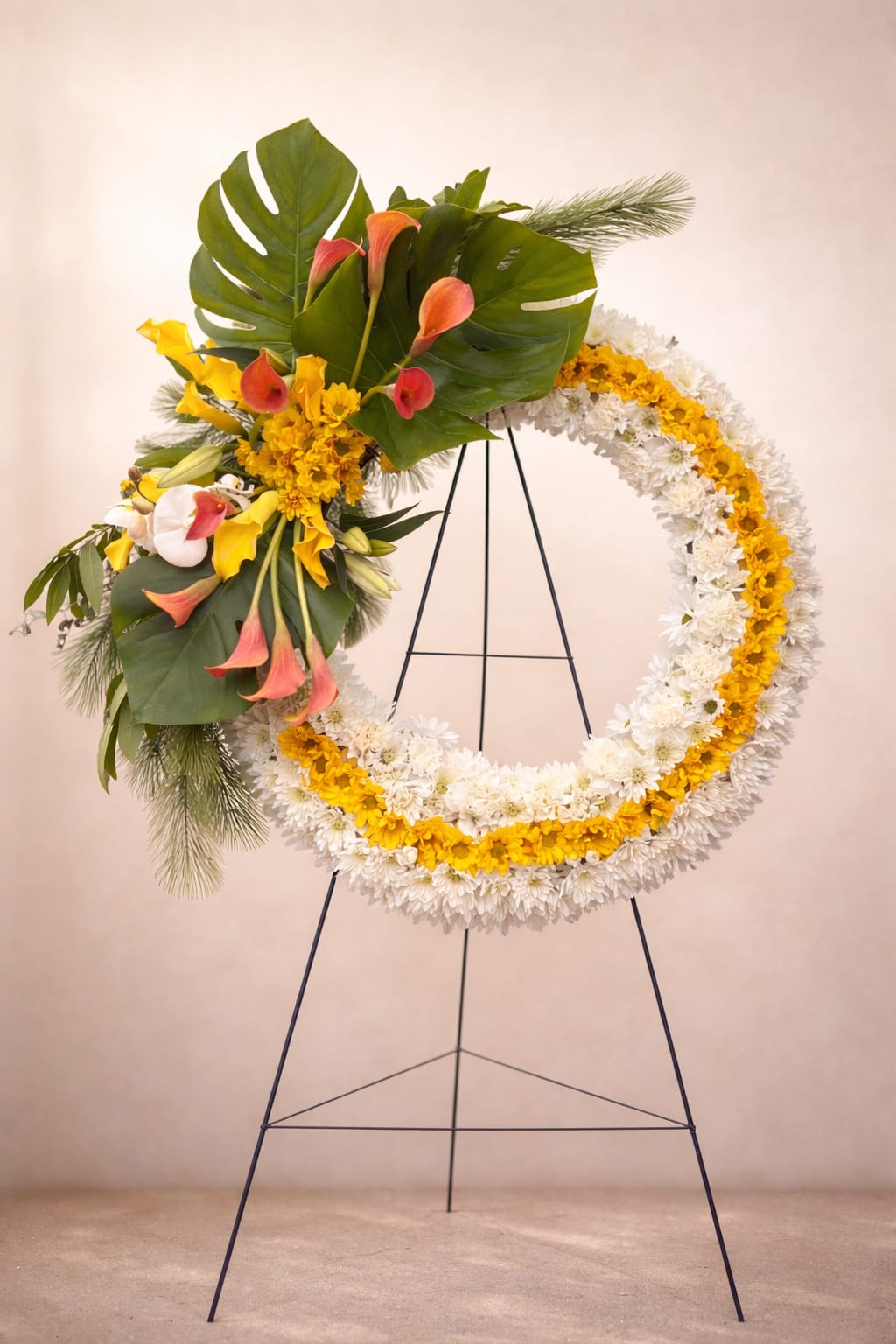 Large white and yellow standing wreath with pink-orange calla lilies and tropical foliage on a metal easel.