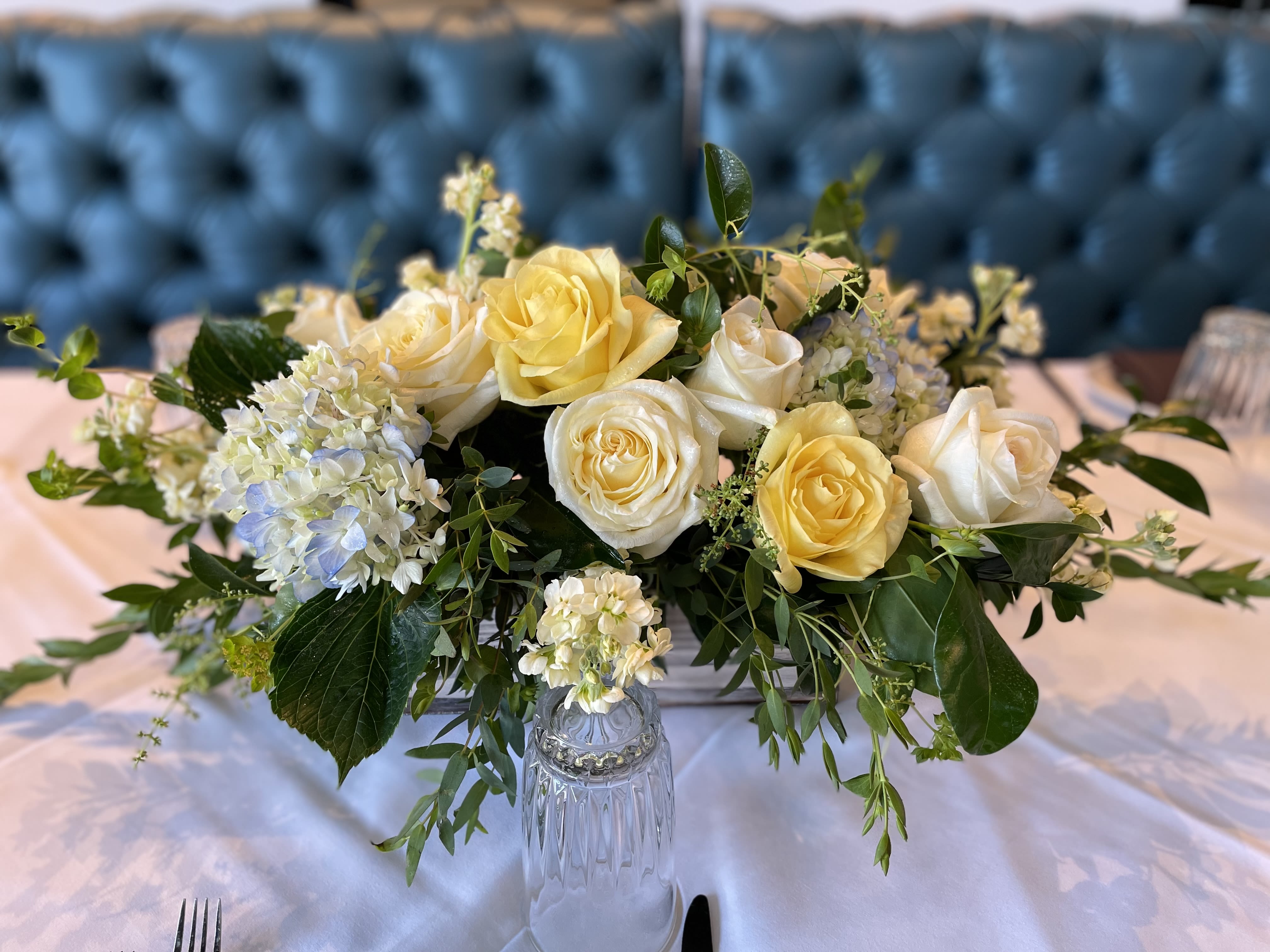Low arrangement of yellow and cream roses with white hydrangeas on a set table.