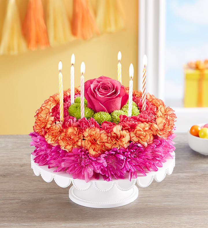 Round floral cake arrangement with pink, orange, and green flowers topped with candles on a white stand