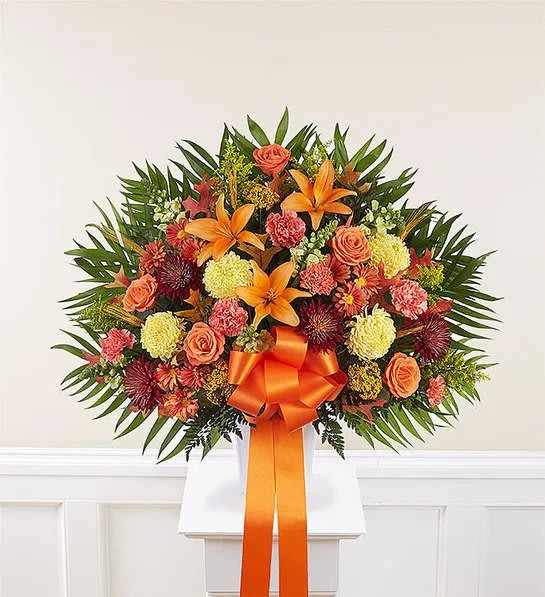 Fan-shaped arrangement of orange lilies, roses, carnations and mums with an orange bow in a white container
