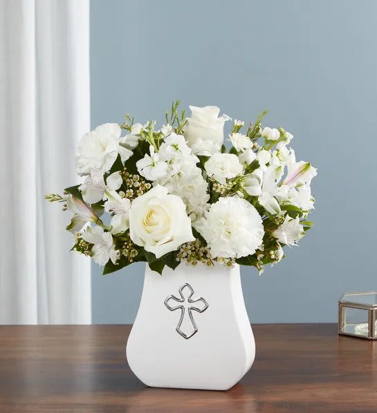 White flower arrangement in a white vase with a silver cross emblem on a wooden table