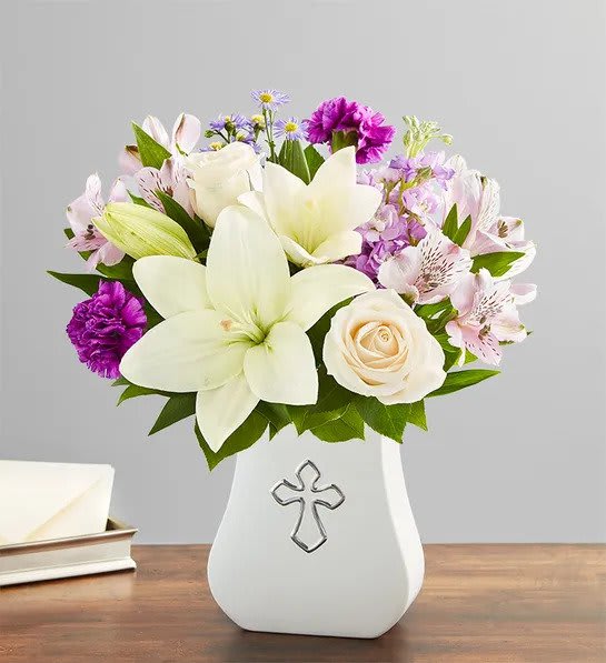 White lilies, roses, and purple flowers in a white vase with a silver cross design.