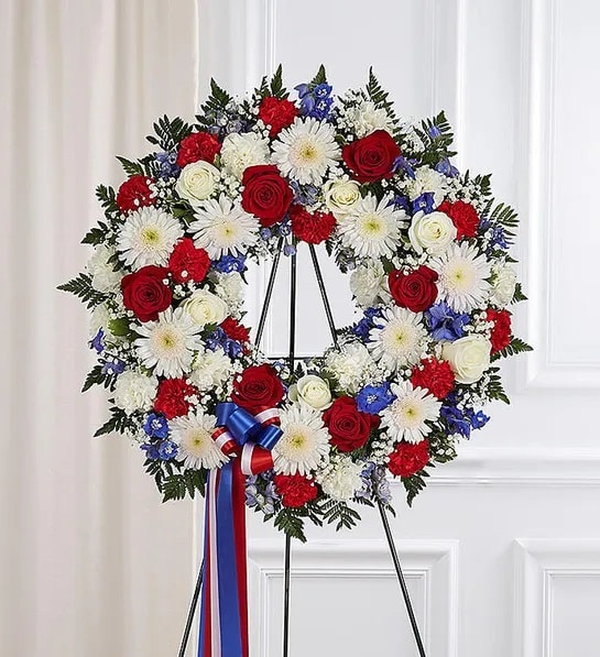 Patriotic standing wreath with red roses, white blooms, and blue flowers on an easel.