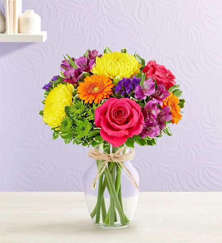 Bright mixed bouquet of pink roses, yellow mums, orange gerberas and purple blooms in a clear glass vase.