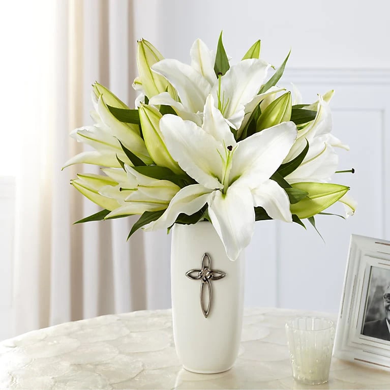 White lilies arranged in a tall white vase with a silver cross emblem on a light table