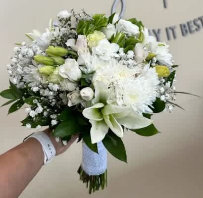 Hand-tied white bouquet with lilies, roses, and mums wrapped in white ribbon, held against a neutral background.