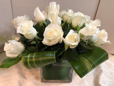 White roses arranged in a square glass vase with broad green leaves